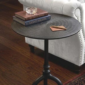Puede incluir: Una mesa auxiliar de metal negro con una superficie texturizada, que contiene libros, un cenicero y un pequeño bloc de notas de cuero con un bolígrafo. La mesa tiene una sola pata central y una base de tres patas. El fondo incluye un sofá blanco y una alfombra estampada.