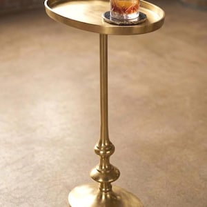 Könnte beinhalten: Ein goldfarbener Beistelltisch mit ovaler Ober- und Unterseite. Der Tisch hat einen schlanken, verzierten Stiel mit dekorativen Details. Ein Glas bernsteinfarbener Flüssigkeit mit Eis steht auf der Tischoberfläche, auf einem dunklen Untersetzer. Der Tisch ist für Getränke konzipiert.