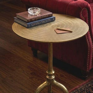 Op de afbeelding: Een ronde bijzettafel van goudkleurig metaal met een textuur van concentrische cirkels op het tafelblad. De tafel heeft een centrale kolomvoet met drie gebogen poten. Twee boeken met de titels Art Deco Jewels en Grand Yachts liggen bovenop.