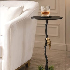 May include: A black side table with a round top, supported by a tree-like stem with a gold-coloured bird. The base features three small planters with green plants. A glass of amber liquid sits on the table.