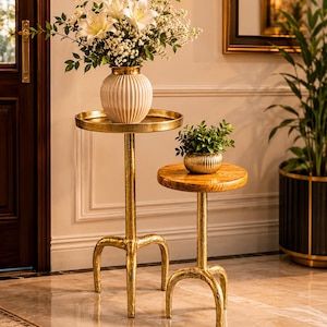 May include: Two gold-toned metal side tables with textured surfaces and three-legged bases. The taller table holds a white ribbed vase with white lilies and greenery, while the shorter table features a small, round, ribbed gold bowl with greenery.