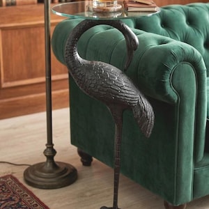 May include: A side table featuring a bronze crane sculpture supporting a clear glass top. The table holds a glass of amber liquid with ice, a coaster, and a notebook. The table is positioned next to a green velvet sofa. The table is a decorative item.