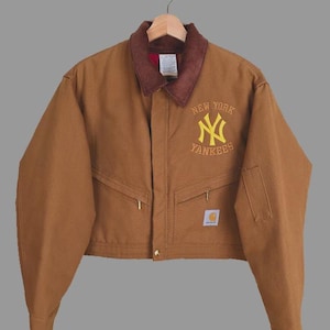 May include: Brown work jacket with a brown corduroy collar and the New York Yankees logo in yellow. The jacket has two front pockets, a small square label, and a button closure. The jacket is hanging on a wooden hanger.
