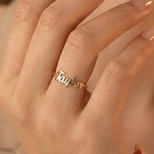 May include: A gold-colored ring with the name "Taylor" in cursive script. The ring is worn on a finger, showcasing the delicate design and personalized touch. The ring is a simple band with the name as the focal point.