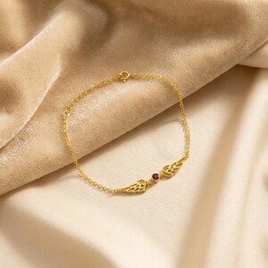 May include: A gold-colored bracelet with a delicate chain and a central design featuring two stylized wings flanking a small, round, red gemstone. The bracelet is displayed on a beige fabric.