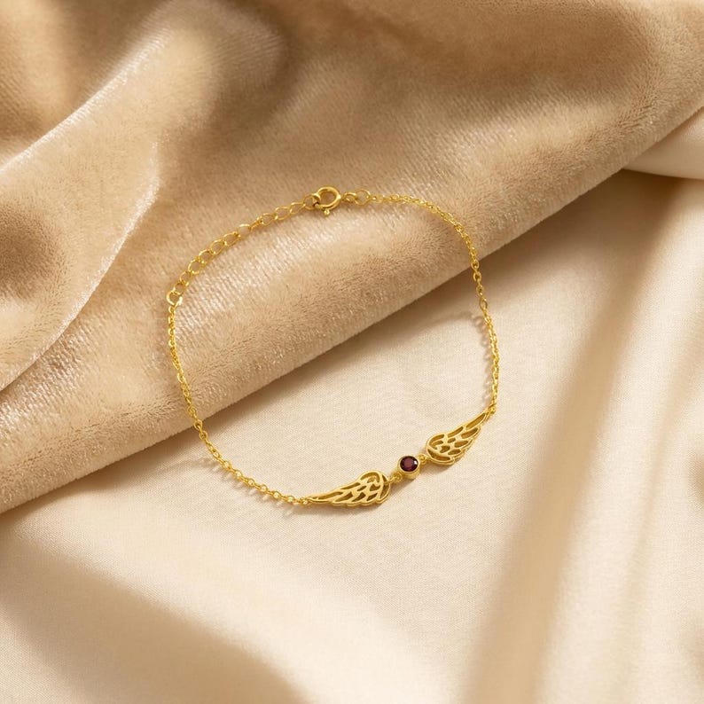 May include: A gold-colored bracelet with a delicate chain and a central design featuring two stylized wings flanking a small, round, red gemstone. The bracelet is displayed on a beige fabric.