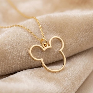 Peut inclure: Un collier doré avec un pendentif en forme de tête de souris de dessin animé célèbre. Le pendentif est un simple contour, et la chaîne est délicate. Le collier est présenté sur un tissu beige doux.