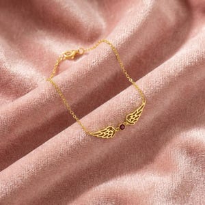 May include: A gold-colored bracelet featuring a pair of stylized wings with intricate detailing. A small, round, red gemstone is set between the wings. The bracelet is displayed on a pink velvet surface.