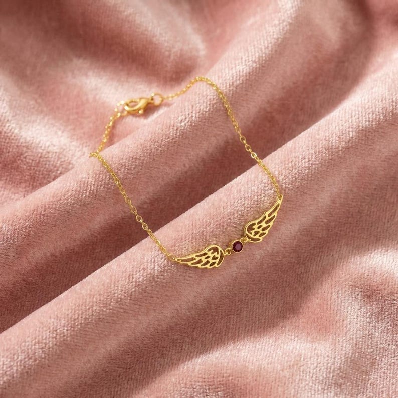 May include: A gold-colored bracelet featuring a pair of stylized wings with intricate detailing. A small, round, red gemstone is set between the wings. The bracelet is displayed on a pink velvet surface.