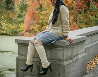 May include: A woman wearing a gray knitted beanie, a cream sweater, a blue skirt, and white knee-high socks sits on a stone wall in a park. The background is a blurry image of fall foliage.