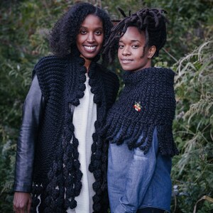 May include: Two people wearing black knitted shawls with fringe. The shawls have a textured, crocheted design. One person is wearing a white shirt and a black leather jacket. The other person is wearing a blue denim shirt and black pants.