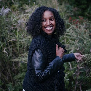 May include: A woman with curly dark hair smiles while wearing a black knitted cardigan with leather sleeves. She is standing in front of a green leafy background.