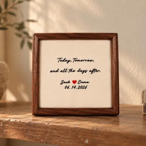 Könnte beinhalten: Ein quadratischer Holzrahmen mit einem cremefarbenen Stoffeinsatz, der den schwarzen Sticktext Today, Tomorrow, and all the days after enthält. Darunter stehen die Namen Jack und Emma, getrennt durch ein rotes Herz, gefolgt von 06.14.2026.