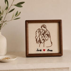 Puede incluir: Un marco de madera cuadrado que contiene un panel de tela color crema con una ilustración bordada en hilo marrón oscuro que muestra a una pareja abrazada. Debajo de la ilustración, los nombres Jack y Rose están bordados en negro con un corazón rojo.