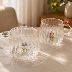 Könnte beinhalten: Zwei Tassen aus klarem Glas mit vertikal gerippter Textur. Jede Tasse hat eine kleine, bunte Glasblume am Henkel und ein Bündel zarter Glasblumen, die aufrecht auf dem inneren Boden der Tasse stehen.