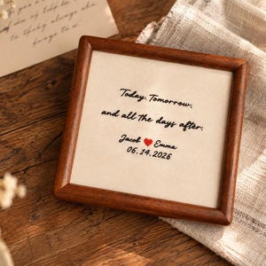 Könnte beinhalten: Ein quadratischer Holzrahmen mit einem cremefarbenen Stoffeinsatz, der den schwarzen Sticktext trägt: Today. Tomorrow, and all the days after. Unter dem Text stehen die Namen Jacob und Emma, getrennt durch ein rotes Herz, sowie das Datum 06.14.2026.