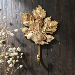 May include: A gold-coloured metal wall hook with a running rabbit design set amongst stylized leaves. The hook is attached to a stem with a rounded end. The background is a dark, textured wooden surface.