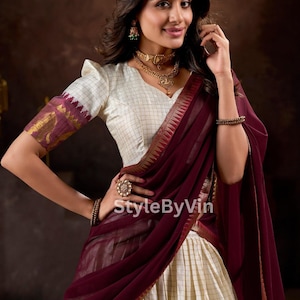 Könnte beinhalten: Eine Frau trägt eine cremefarbene, karierte Bluse mit bordeauxfarbenen Akzenten und einen passenden drapierten Schal. Sie ist mit Goldschmuck geschmückt, darunter eine Halskette, Ohrringe und ein Kopfschmuck. Das Outfit wird durch einen cremefarbenen Rock ergänzt.