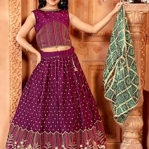 May include: A young girl in a traditional Indian outfit. The ensemble includes a sleeveless, purple crop top with striped and dotted details, a matching skirt, and a green patterned scarf. The skirt has a detailed floral border.