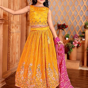 Könnte beinhalten: Ein ärmelloses, senfgelbes Kleid mit aufwendiger Stickerei und einem passenden Rock. Der Rock hat ein Pfauenmuster. Ein pinkfarbener Schal und ein Armband mit Muster vervollständigen das Outfit. Das Mädchen trägt eine Sonnenbrille und ein Stirnband.