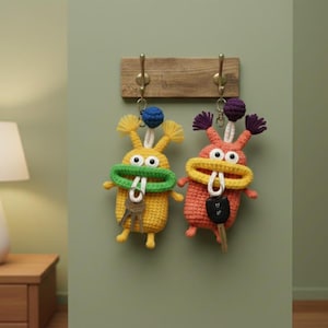 May include: Two handmade crochet keychains shaped like whimsical creatures, one yellow with a green mouth and the other orange with a yellow mouth. Each has a key attached and hangs from a wooden rack with brass hooks.