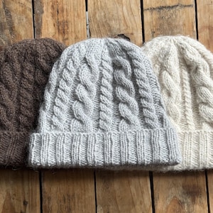 May include: Three knit beanies featuring a cable knit pattern and a ribbed cuff. The beanies are arranged side by side in dark brown, light gray, and off-white colors, showcasing a thick, textured wool-like yarn construction.