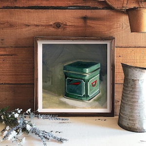 May include: A framed painting of a green tin box with red and white lettering on a dark background. The box is sitting on a table with a light brown surface.