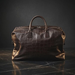 May include: A dark brown travel bag with a textured crocodile-embossed pattern. The bag features two top handles, a horizontal zip closure across the top, and metallic hardware attachments on the sides for a shoulder strap.