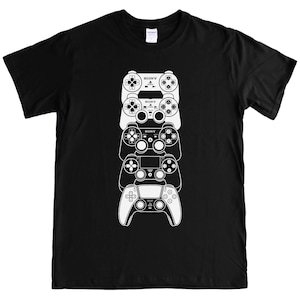 May include: Black t-shirt with a white graphic of stacked video game controllers. The controllers include the Sony logo and various button layouts, showing a progression of designs. The shirt is made of a soft material.