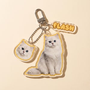 May include: A keychain featuring a fluffy white cat with blue eyes. The keychain has three acrylic charms: a full cat, a cat head, and a charm that says "FLASH" in yellow letters. The keychain is attached to a silver metal clasp.