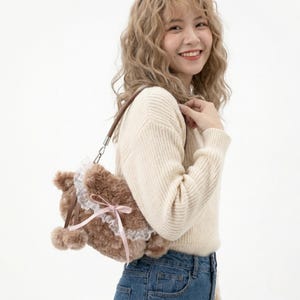 May include: A brown teddy bear-shaped handbag with a white lace trim and a pink ribbon. The bag has a brown leather strap and two small pom-poms. The person is wearing a cream-colored sweater and blue jeans. The bag is a cute accessory.