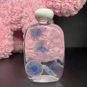 May include: A clear glass bottle with a white stopper, containing several blue and white jellyfish. The bottle has a rounded shape and is filled with a pink liquid. The jellyfish have detailed tentacles and are scattered throughout the bottle.
