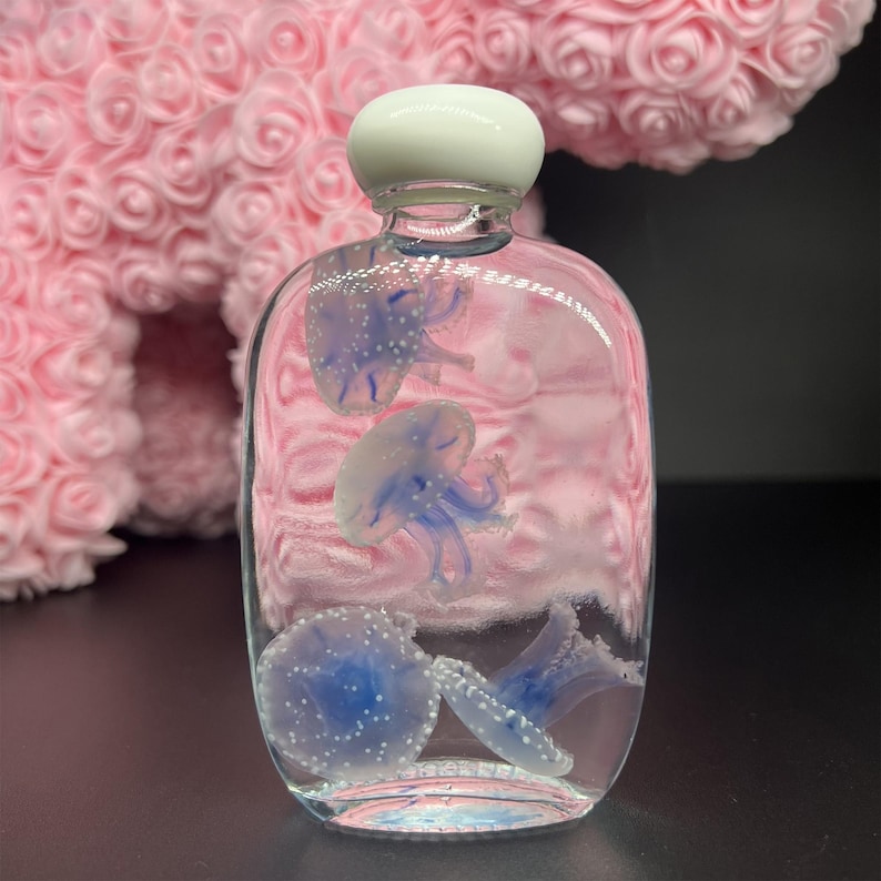 May include: A clear glass bottle with a white stopper, containing several blue and white jellyfish. The bottle has a rounded shape and is filled with a pink liquid. The jellyfish have detailed tentacles and are scattered throughout the bottle.