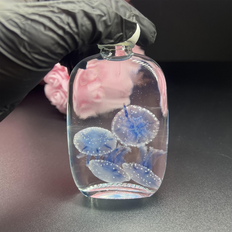 May include: A clear glass bottle with a rounded shape, containing several blue and white jellyfish. The jellyfish have a detailed design. The bottle has a clear glass stopper.