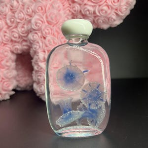 May include: A clear glass bottle with a white stopper, featuring blue and white jellyfish designs inside. The bottle has a rounded shape and is filled with a clear liquid. The jellyfish have a detailed, dotted pattern.