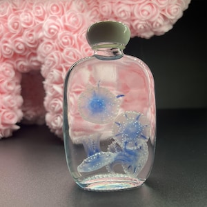 May include: A clear, rectangular glass bottle with a light gray cap, containing blue and white jellyfish designs. The bottle is filled with a clear liquid, and the jellyfish have detailed, delicate features.
