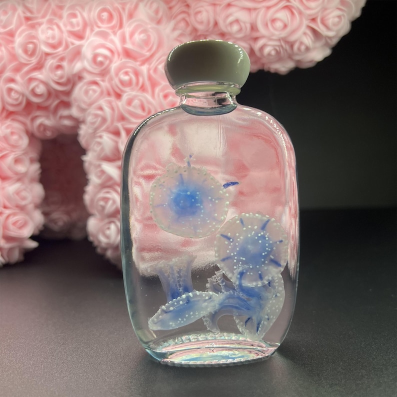 May include: A clear, rectangular glass bottle with a light gray cap, containing blue and white jellyfish designs. The bottle is filled with a clear liquid, and the jellyfish have detailed, delicate features.