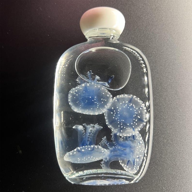 May include: A clear glass bottle with a white stopper, containing blue jellyfish with white spots. The bottle has a unique shape and a bubble-like design element. The jellyfish are suspended in the liquid.
