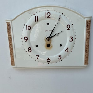 May include: Cream-coloured vintage wall clock with a rectangular shape and arched top. The clock face has black hands, red numerals, and the brand name "INGERSOLL". The sides feature a brown patterned design.