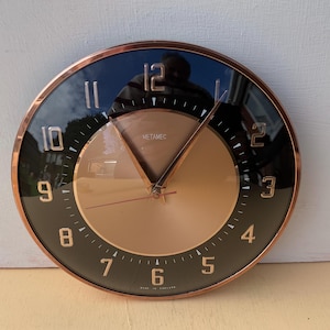May include: A round wall clock with a copper-colored bezel and a black and gold face. The clock has gold-colored hands and numerals. The text "METAMEC" is visible on the face, and "MADE IN ENGLAND" is at the bottom.
