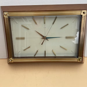 以下が含まれることがあります： 木製のフレームと金色のトリムが付いた長方形の壁掛け時計。文字盤はオフホワイトで、金と青の針と金色の時間マーカーが付いています。時計はミッドセンチュリーモダンデザインです。