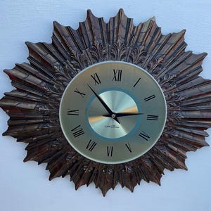 Peut inclure: Une grande horloge murale ronde de couleur or avec des chiffres romains. Le cadran de l'horloge est entouré d'un cadre décoratif en forme de soleil éclatant avec une finition métallique brune.
