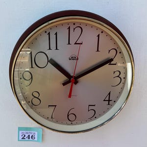 May include: A round wall clock with a brown frame, gold trim, and a light-colored face. The clock features black numerals, black hands, and a red second hand. The brand name "Smiths Timeco" is printed on the face. A small blue tag with the numbers "AD397616" and "246" is in the lower left corner.