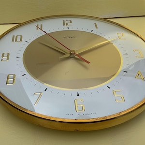 May include: A circular wall clock with a gold-coloured frame, white face, and gold numerals. The clock features a gold centre and a red second hand. The face is marked with "METAMEC" and "MADE IN ENGLAND".