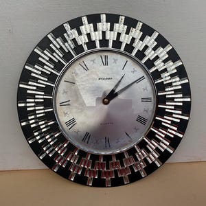 May include: A circular wall clock with a black surround and a silver-toned face. The clock displays Roman numerals and the word "STAVGER". The frame is adorned with mirrored rectangular details. The clock hands are black.