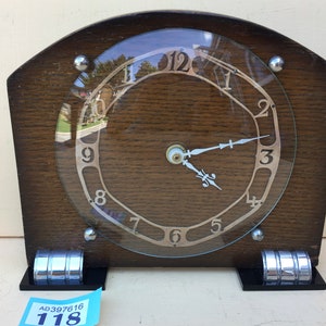 Puede incluir: Un reloj vintage de madera con una cara de vidrio y detalles plateados. El reloj tiene marcas de números romanos y una aguja plateada. El reloj está sentado sobre una base negra con detalles plateados. El reloj está etiquetado con el número "118".