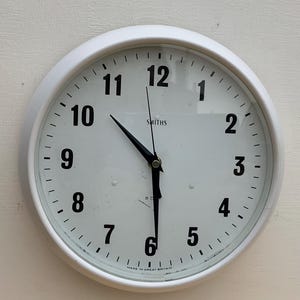 Puede incluir: Reloj de pared analógico blanco con agujas negras y números negros. La esfera del reloj tiene el texto "SMITHS" y "MADE IN GREAT BRITAIN".