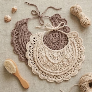 May include: Two handmade crochet baby bibs in beige and brown, with scalloped edges and tie closures. A wooden baby brush, seashells, and yarn are also visible.