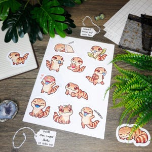 May include: A sheet of stickers featuring cartoon blue-tongue skinks in various poses. The stickers are in shades of brown and orange with blue tongues. The text "Sweet Blue Tongue Skinks (12 Sticker Set)" is on a tag.