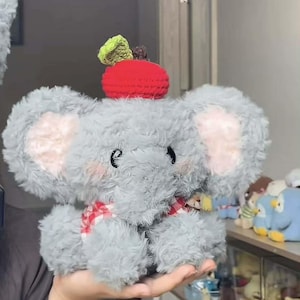 Puede incluir: Un peluche de elefante gris con orejas grandes y esponjosas y una bufanda a cuadros rojos y blancos. El elefante tiene una manzana roja de ganchillo en la cabeza con una hoja verde. El juguete es suave y tierno.